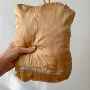 Puede incluir: Una bolsa plegada, de color dorado, similar al sat&eacute;n, con un cierre de cremallera plateado en la parte inferior. La bolsa tiene un lazo en la parte superior para llevarla. La bolsa est&aacute; sujeta en la mano de una persona.