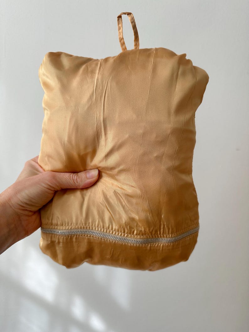 Puede incluir: Una bolsa plegada, de color dorado, similar al sat&eacute;n, con un cierre de cremallera plateado en la parte inferior. La bolsa tiene un lazo en la parte superior para llevarla. La bolsa est&aacute; sujeta en la mano de una persona.