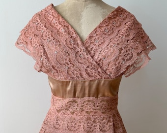 Puede incluir: Un vestido vintage de encaje rosa con una cintura de satén y un escote en V. El vestido tiene una falda de capas y mangas cortas.