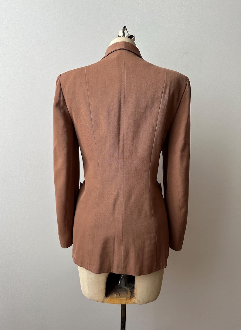 May include: A brown blazer displayed on a mannequin. The tailored jacket features a notched lapel, two front pockets, and a fitted silhouette. The back view highlights the structured design and the warm, earthy tone of the fabric.