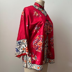 May include: A red silk Chinese-style jacket with floral embroidery. The jacket has a mandarin collar and a front closure with buttons. The sleeves are three-quarter length and have a decorative trim.