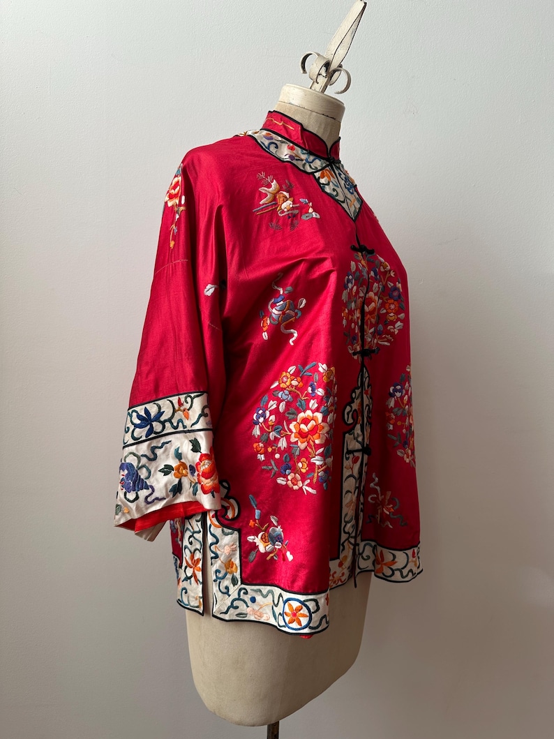 May include: A red silk Chinese-style jacket with floral embroidery. The jacket has a mandarin collar and a front closure with buttons. The sleeves are three-quarter length and have a decorative trim.