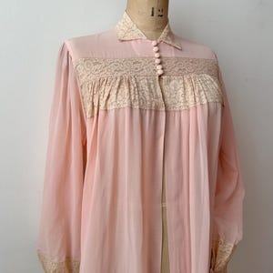 Op de afbeelding: Een vintage, lichtroze badjas met lange mouwen en een kraag. De badjas heeft delicate kanten details over de borst en bij de manchetten. Het is aan de voorkant dichtgeknoopt en lijkt gemaakt van een lichte, vloeiende stof.