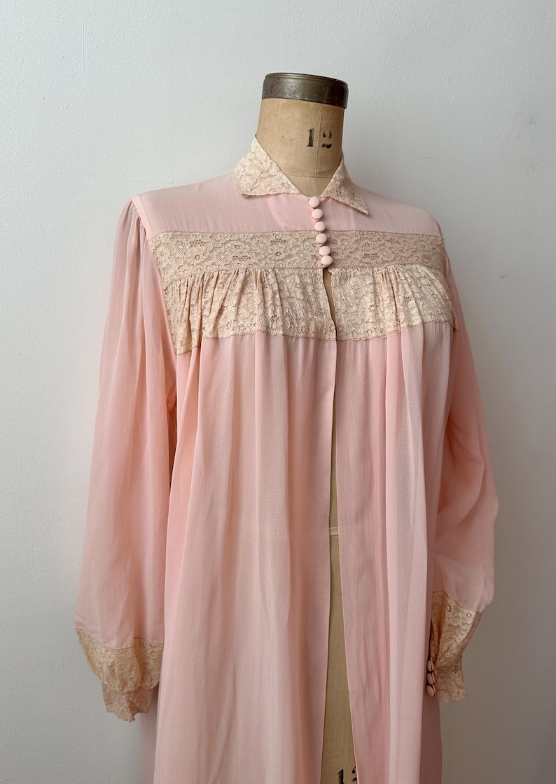 Op de afbeelding: Een vintage, lichtroze badjas met lange mouwen en een kraag. De badjas heeft delicate kanten details over de borst en bij de manchetten. Het is aan de voorkant dichtgeknoopt en lijkt gemaakt van een lichte, vloeiende stof.
