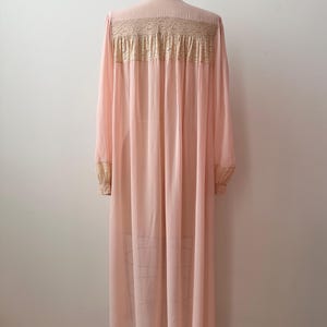 Op de afbeelding: Een vintage, lichtroze nachtjapon met lange mouwen en een hoge kraag. De japon heeft delicate kanten details over de bovenrug en op de manchetten. De stof drapeert sierlijk en toont een klassiek, elegant ontwerp.