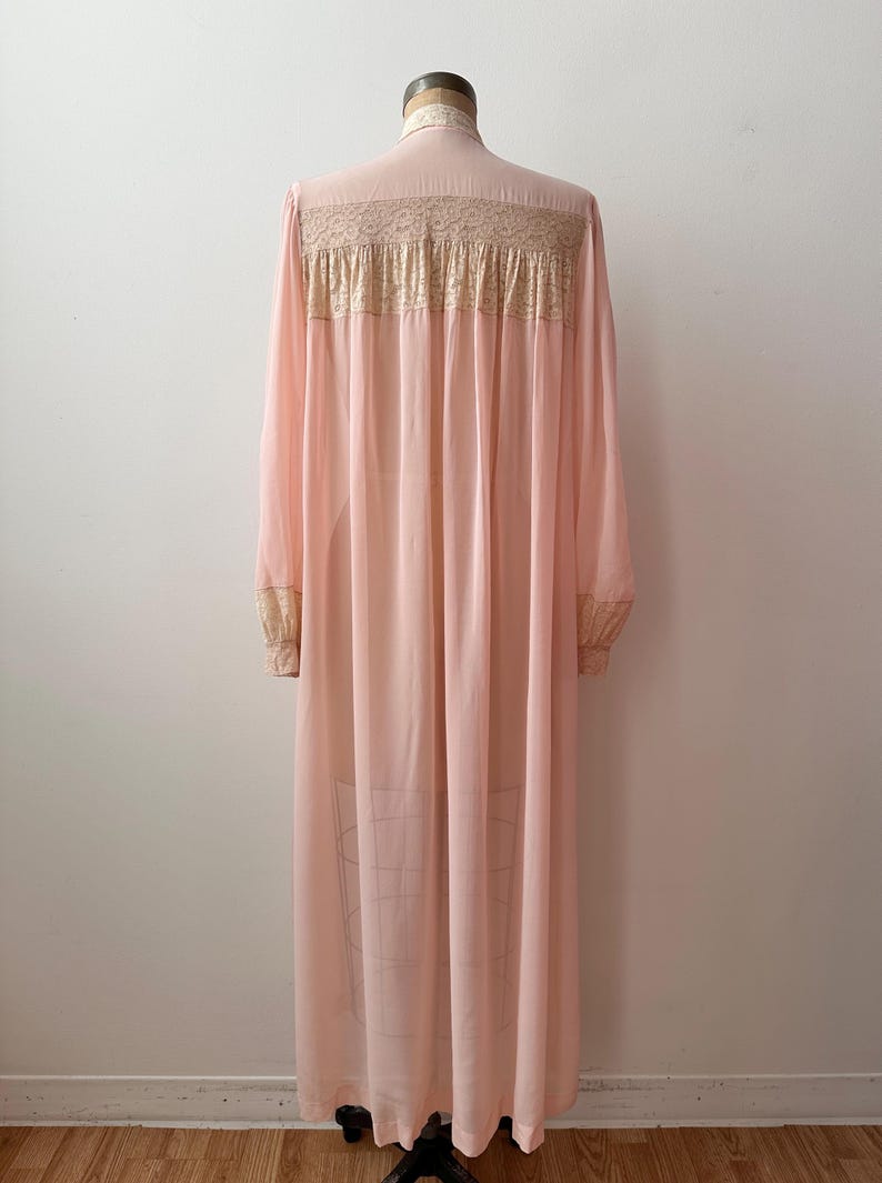 Op de afbeelding: Een vintage, lichtroze nachtjapon met lange mouwen en een hoge kraag. De japon heeft delicate kanten details over de bovenrug en op de manchetten. De stof drapeert sierlijk en toont een klassiek, elegant ontwerp.
