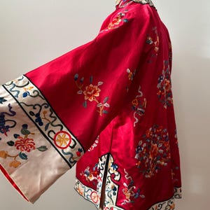 May include: A red silk Chinese jacket with floral embroidery and a white trim. The jacket has a mandarin collar and long sleeves.