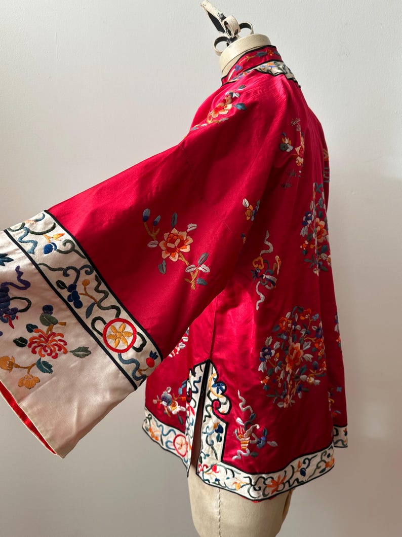 May include: A red silk Chinese jacket with floral embroidery and a white trim. The jacket has a mandarin collar and long sleeves.