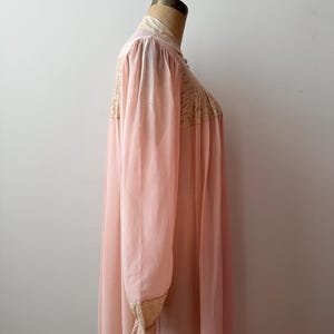 Op de afbeelding: Een vintage, lichtroze nachtjapon met lange mouwen en een hoge kraag. De nachtjapon heeft delicate kanten details op het lijfje, de kraag en de manchetten. De stof lijkt een lichtgewicht, vloeiend materiaal te zijn.