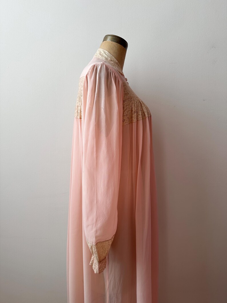 Op de afbeelding: Een vintage, lichtroze nachtjapon met lange mouwen en een hoge kraag. De nachtjapon heeft delicate kanten details op het lijfje, de kraag en de manchetten. De stof lijkt een lichtgewicht, vloeiend materiaal te zijn.