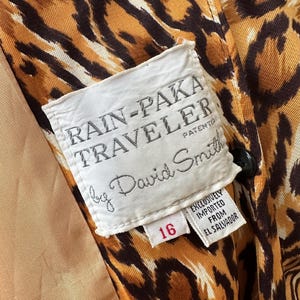 Puede incluir: Primer plano del interior de un impermeable vintage con estampado de leopardo. El forro es de color beige claro. Una etiqueta blanca dice "RAIN-PAKA TRAVELER by David Smith" con el n&uacute;mero "16" en rojo. El abrigo se importa exclusivamente de El Salvador.