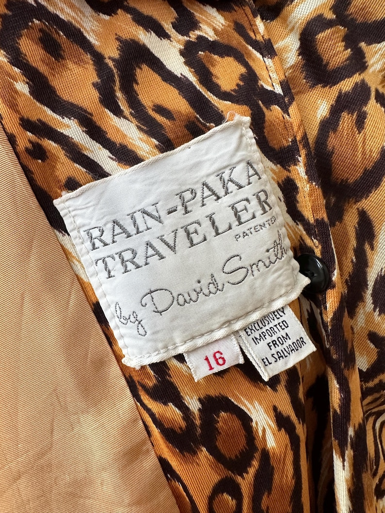 Puede incluir: Primer plano del interior de un impermeable vintage con estampado de leopardo. El forro es de color beige claro. Una etiqueta blanca dice "RAIN-PAKA TRAVELER by David Smith" con el n&uacute;mero "16" en rojo. El abrigo se importa exclusivamente de El Salvador.