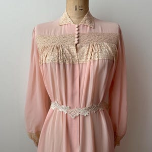 Op de afbeelding: Een vintage roze badjas met lange mouwen en een kraag. De badjas heeft een cr&egrave;mekleurig kanten paneel over de borst en een bijpassende kanten riem. De badjas wordt op een mannequin gepresenteerd.