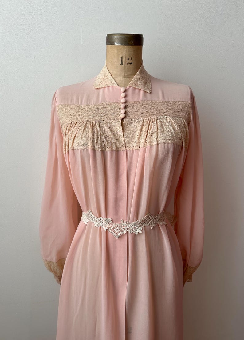 Op de afbeelding: Een vintage roze badjas met lange mouwen en een kraag. De badjas heeft een cr&egrave;mekleurig kanten paneel over de borst en een bijpassende kanten riem. De badjas wordt op een mannequin gepresenteerd.
