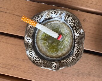 May include: A ceramic ashtray with a speckled brown and black rim, holding a beaded cigarette. The cigarette has a white, orange, and red design. The ashtray's centre has a green, textured appearance.
