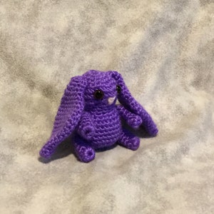 May include: A small, purple crocheted rabbit toy featuring long, floppy ears, a round body, and two dark, glossy plastic eyes. The stuffed animal is crafted with a visible yarn stitch pattern and includes a small, light-colored nose detail.