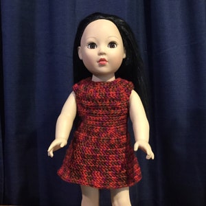 May include: A doll wearing a red, brown, and purple crocheted dress. The dress has a sleeveless design and a flared skirt.