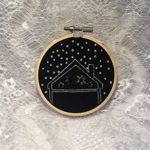 May include: A circular wooden embroidery hoop framing black fabric with white thread stitching depicting a house outline and star-like shapes. Small white beads are scattered across the upper portion of the fabric to represent falling snow.