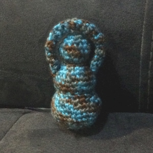 May include: A crocheted doll with a brown and blue striped pattern. The doll has a round head and a long, thin body.