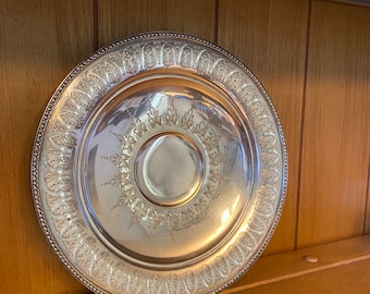 Puede incluir: Un plato de servir de plata con un diseño ornamentado. El plato tiene un borde elevado y un pozo central. El diseño presenta un patrón repetitivo de hojas y espirales.