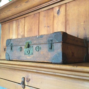 May include: A vintage wooden suitcase with metal clasps and hinges. The suitcase is brown and has a worn, aged look. It is sitting on a wooden shelf.