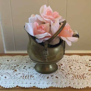 May include: A vintage brass coal scuttle filled with pink and white fabric roses. The scuttle has a handle and sits on a wooden surface with a white lace doily. The background is a light-colored wall.