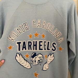 Op de afbeelding: Lichtblauw sweatshirt met een witte en donkerblauwe afbeelding van het logo van de North Carolina Tarheels. Het logo toont een cartoon mascotte met een basketbal en sterren.