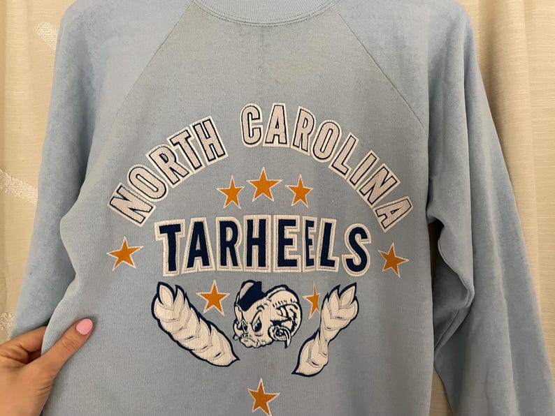 Op de afbeelding: Lichtblauw sweatshirt met een witte en donkerblauwe afbeelding van het logo van de North Carolina Tarheels. Het logo toont een cartoon mascotte met een basketbal en sterren.