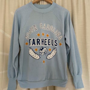 Op de afbeelding: Lichtblauw sweatshirt met een vintage North Carolina Tar Heels logo in oranje, blauw en wit. Het logo toont een mascotte, sterren en de woorden "North Carolina Tar Heels".