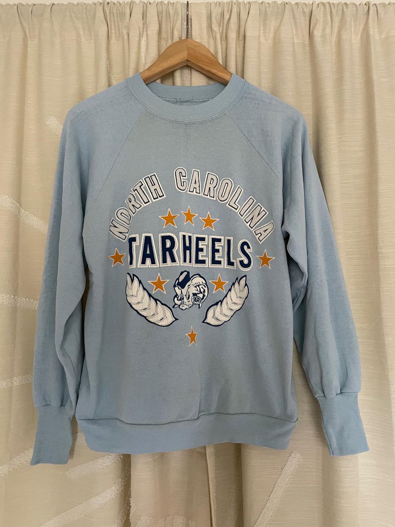 Op de afbeelding: Lichtblauw sweatshirt met een vintage North Carolina Tar Heels logo in oranje, blauw en wit. Het logo toont een mascotte, sterren en de woorden "North Carolina Tar Heels".