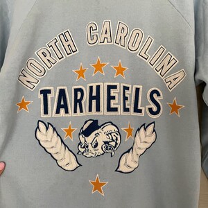 Op de afbeelding: Lichtblauw sweatshirt met de tekst "North Carolina Tarheels" in witte en donkerblauwe letters. Het sweatshirt heeft een cartoon mascotte met een stervormig ontwerp.