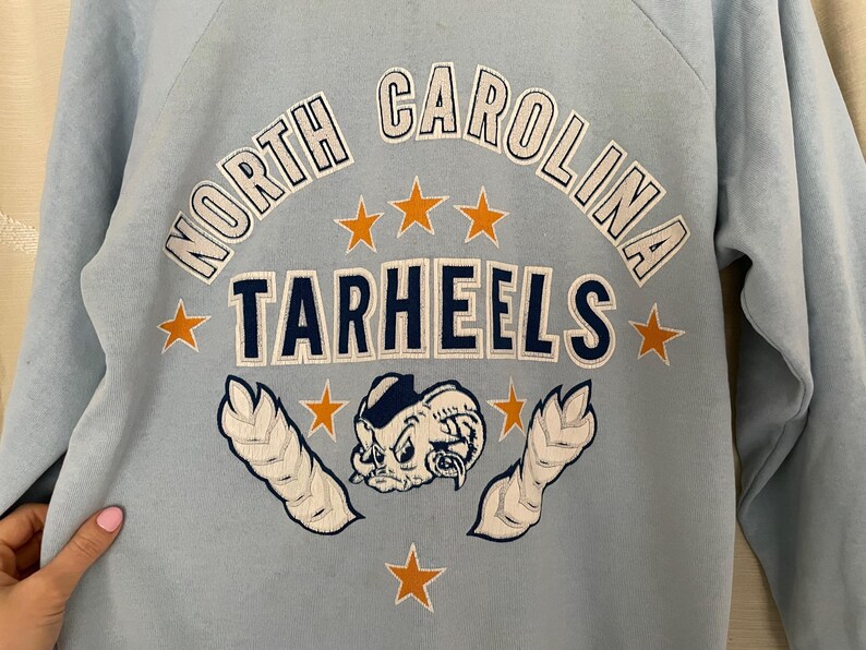 Op de afbeelding: Lichtblauw sweatshirt met de tekst "North Carolina Tarheels" in witte en donkerblauwe letters. Het sweatshirt heeft een cartoon mascotte met een stervormig ontwerp.
