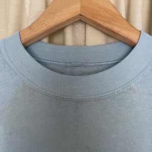 Op de afbeelding: Een lichtblauw sweatshirt met een wit en blauw geborduurd logo. Het sweatshirt hangt aan een houten hanger.