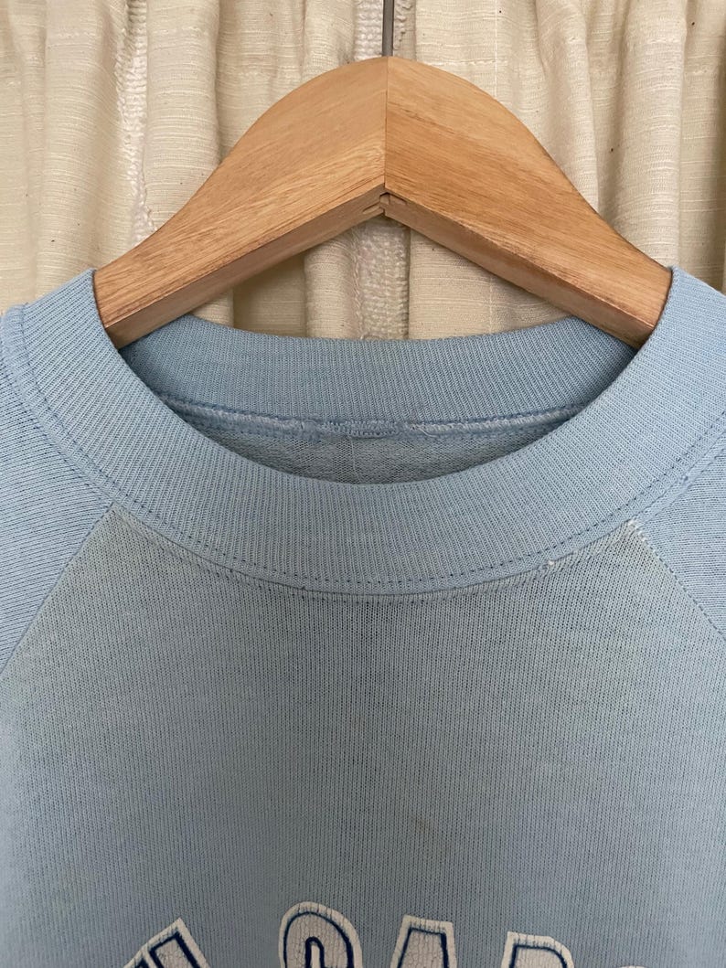 Op de afbeelding: Een lichtblauw sweatshirt met een wit en blauw geborduurd logo. Het sweatshirt hangt aan een houten hanger.
