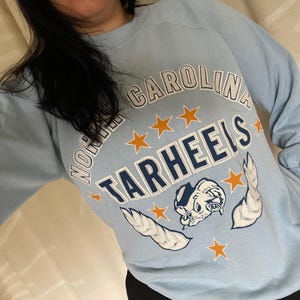 Op de afbeelding: Lichtblauw sweatshirt met de tekst "North Carolina Tarheels" in witte en oranje letters. Het sweatshirt heeft een cartoon mascotte met vleugels en sterren.