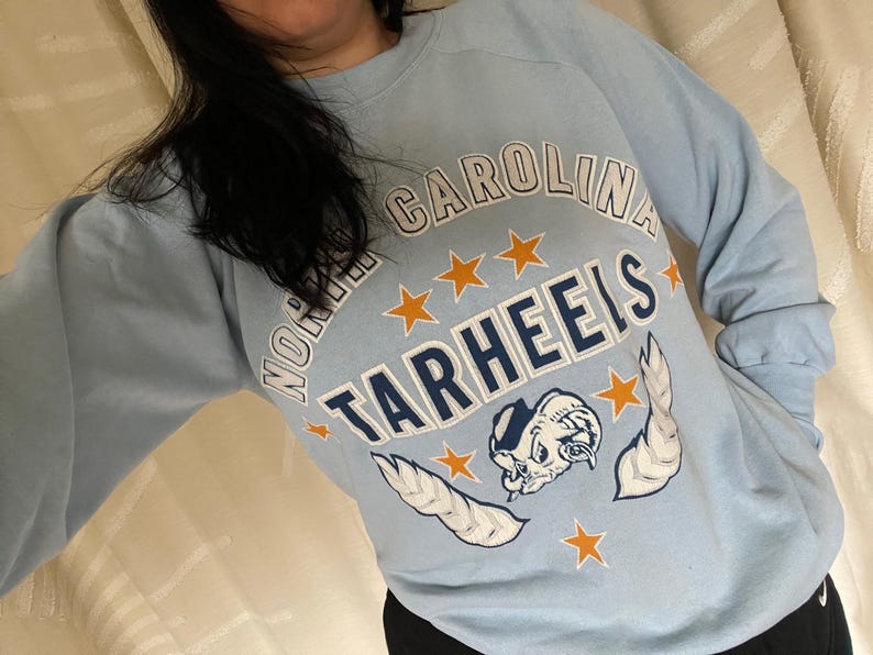 Op de afbeelding: Lichtblauw sweatshirt met de tekst "North Carolina Tarheels" in witte en oranje letters. Het sweatshirt heeft een cartoon mascotte met vleugels en sterren.