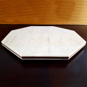 May include: An octagonal marble serving board with a white surface and brown trim. The board is set on a dark brown surface, with a wooden background.