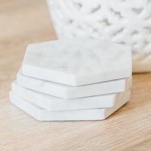 Set of handcrafted Carrara marble hexagon coasters with soft grey veining, displayed on a light wood surface next to a white ceramic vase. Elegant modern drink coasters with a polished or honed finish, available in sets of 4, 6, or 8.