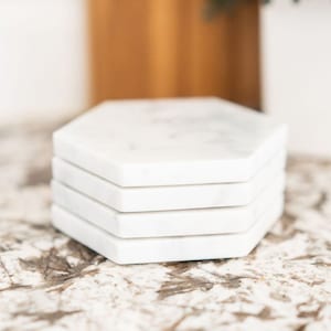 May include: A set of four white marble hexagon-shaped coasters. The coasters are stacked on top of each other and have a subtle marbled pattern.