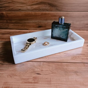 Natural marble rectangle tray with a gold watch, rings, and cologne arranged on top, showing the tray in use as a valet catchall for everyday items on a nightstand or bathroom counter.