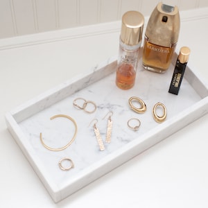 Carrara marble tray holding gold jewelry and perfume bottles on a white surface. The tray's white marble with gray veining provides an elegant display for accessories.
