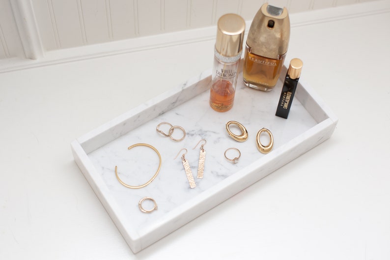 Carrara marble tray holding gold jewelry and perfume bottles on a white surface. The tray's white marble with gray veining provides an elegant display for accessories.