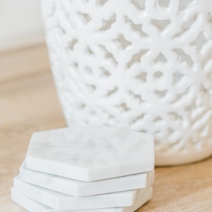 May include: A set of four white marble hexagonal coasters. The coasters are stacked on top of each other and are sitting on a light brown wooden surface.