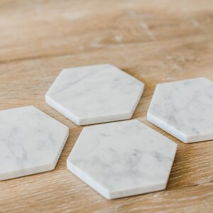 Set of four handcrafted Carrara marble hexagon coasters with soft grey veining, displayed on a natural wood surface. Modern stone drink coasters with polished or honed finish, perfect for protecting tabletops in kitchen or living room settings.