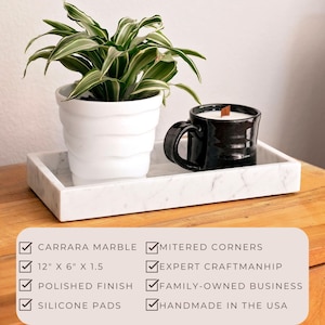Carrara marble tray on a wooden surface holding a potted plant and a black ceramic candle. Features include Carrara marble, mitered corners, polished finish, silicone pads, dimensions 12" x 6" x 1.5", expert craftsmanship, handmade in the USA.