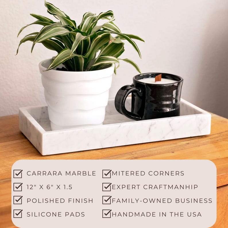Carrara marble tray on a wooden surface holding a potted plant and a black ceramic candle. Features include Carrara marble, mitered corners, polished finish, silicone pads, dimensions 12" x 6" x 1.5", expert craftsmanship, handmade in the USA.