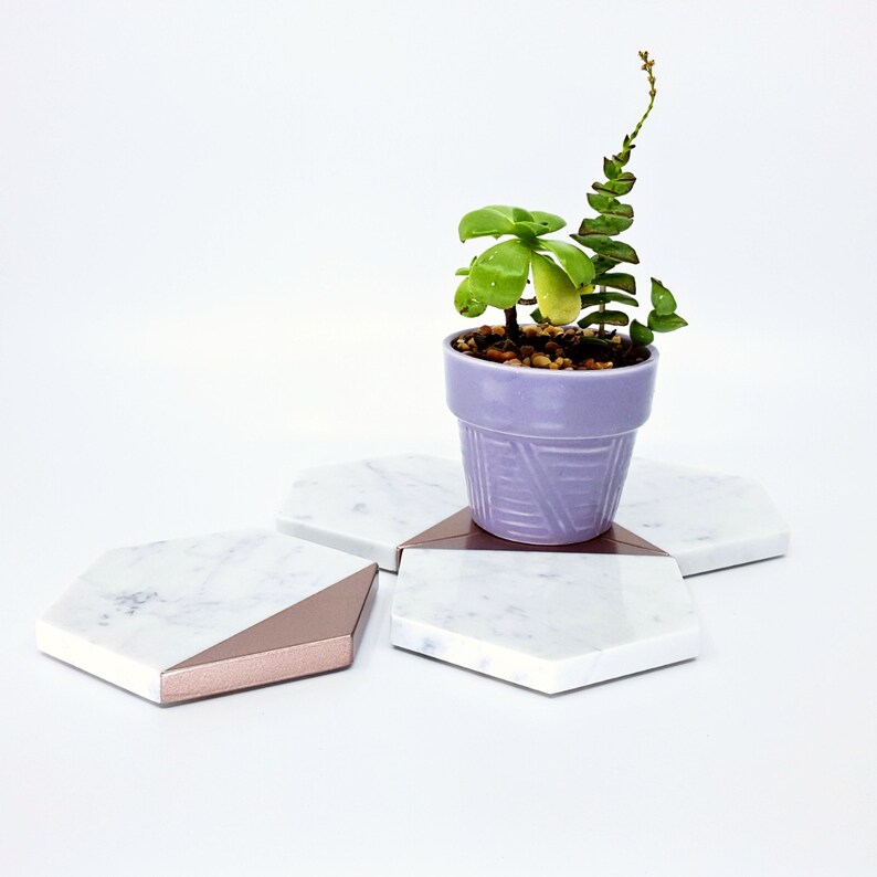 May include: Three hexagonal marble coasters with a rose gold trim. The coasters are arranged in a star shape with a small potted succulent plant in the center.