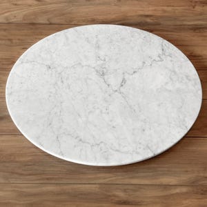 May include: A circular white marble slab featuring natural grey veining throughout the surface. The stone has a smooth, polished finish and a consistent rounded edge, showcasing the organic patterns and textures inherent in the natural material.