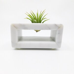 May include: A white marble planter with a rectangular opening, designed to hold an air plant. The air plant is green and has long, thin leaves.