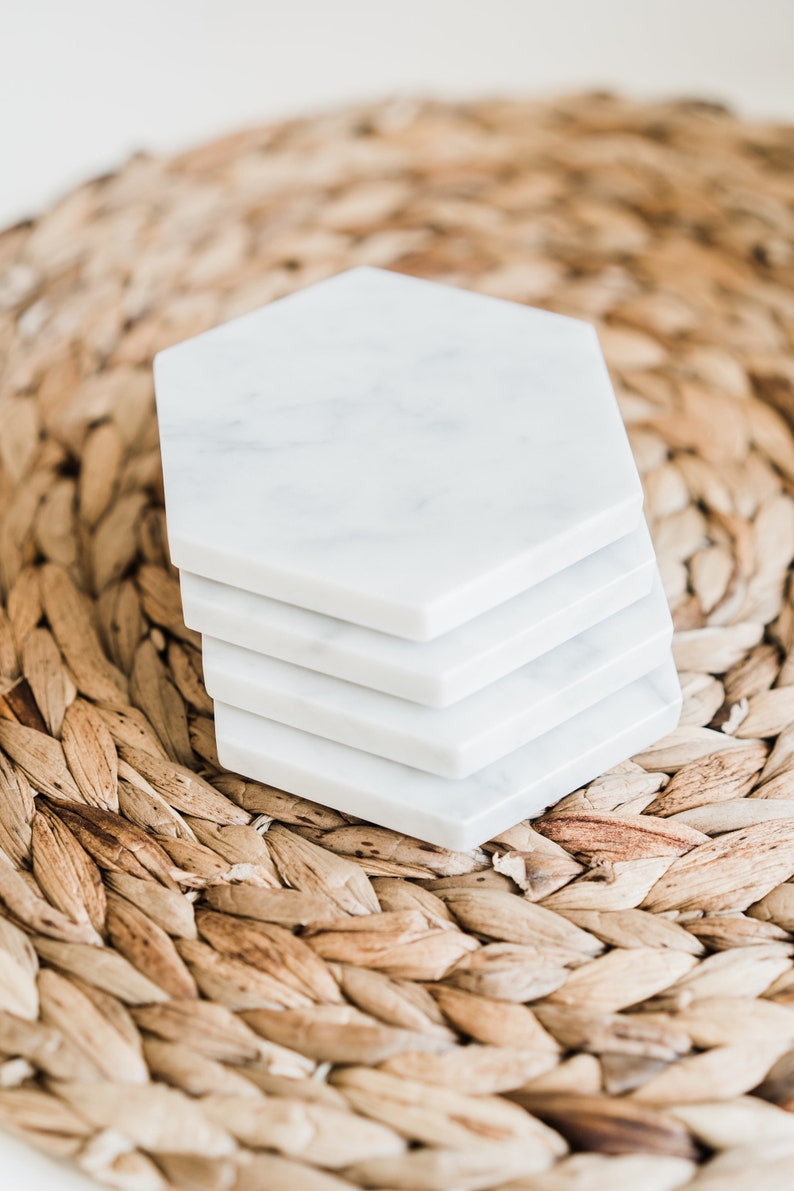 White Carrara marble hexagon coasters with subtle grey veining, styled on a woven natural fiber placemat. Handmade stone coasters with a modern look, ideal for kitchen, dining, or coffee table decor.